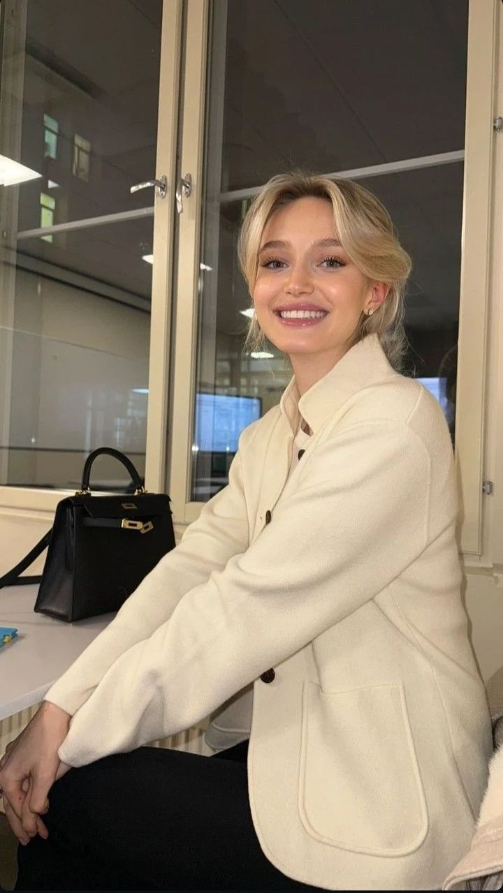 Smiling woman in a cream jacket seated beside a window